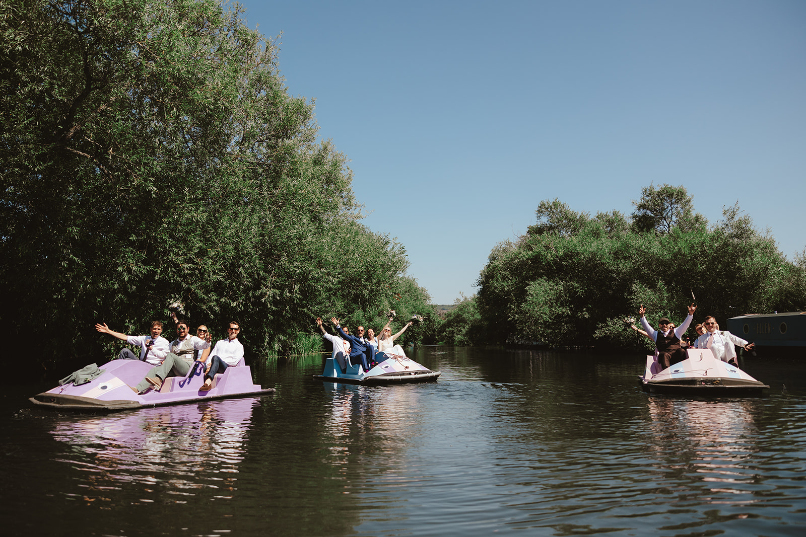 Bride, groom and wedding guests waving from pedalos on a river on a sunny wedding day